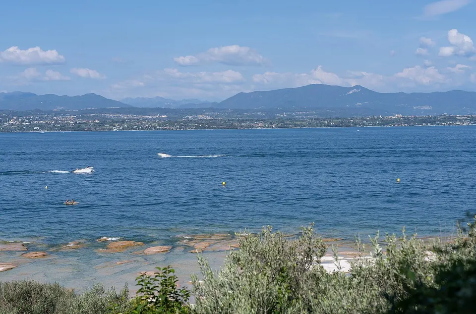Boote im klaren Wasser des Gardasees bei Sirmione, am Ufer stehen Olivenbäume